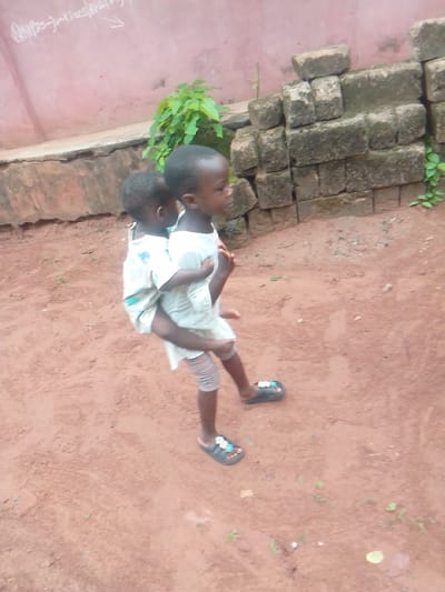 In Alayi, Nigeria, a child wearing sandals is carrying another child on their back. They are standing on a dirt surface. Behind them is a low wall, a stack of bricks, and a green plant. Graffiti is visible on a pink wall in the background.