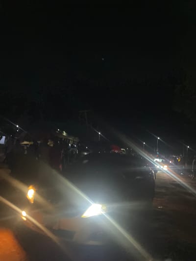 The image depicts a nighttime scene in Sauka, Nigeria, with a gathering of people and motorcycles. The lighting is dim, with the primary illumination coming from bright, directional lights, possibly from vehicles or lamps, creating strong lens flares that obscure some details. Several motorcycles are visible, suggesting a mode of transportation or part of the event. Figures of people are present, though their features are not clearly discernible due to the darkness and the quality of the light. Some individuals appear to be standing or moving around the motorcycles. The scene appears to be outdoors, possibly on a road or open area. The overall impression is of a dynamic, possibly social or economic activity taking place after dark. The time of day is clearly night. There are no visible text elements.
