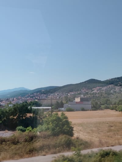 The image shows a view of the city of Lagyna, Greece, with a residential area nestled on a hillside. The foreground features dry, brown grass and shrubs, with some trees and a paved road. In the mid-ground, there is a large, flat-topped building with a red sign that appears to say "Alouystem". Beyond this building, the city of Lagyna stretches up the hill, characterized by numerous houses with red-tiled roofs. The background consists of green, tree-covered hills under a clear blue sky. The overall scene depicts a sunny day in a semi-rural setting within a Greek town.