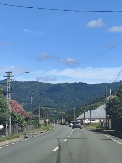 The image depicts a view from a vehicle traveling on a paved road through the village of Leordina, Romania. The road is bordered by vegetation and a few houses with reddish-brown roofs. On the left side of the road, a utility pole with multiple wires and streetlights is visible. Further down the road, two cars are seen in motion. The backdrop is dominated by a densely forested mountain range under a bright blue sky with scattered clouds.