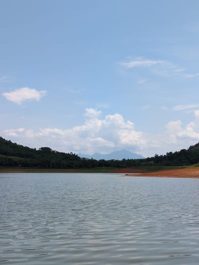This is a wide-angle, eye-level, full shot of a calm body of water, likely a lake or reservoir, with a distinct undulating surface. The water occupies the lower half of the image. The shoreline to the right is characterized by exposed reddish-brown earth and scattered vegetation. Beyond the water, a densely forested hillside slopes upwards, transitioning into a backdrop of distant, hazy mountains under a bright blue sky dotted with white, fluffy clouds. The scene evokes a natural, serene landscape, devoid of any human presence or activity. The location is identified as Ngoc Lac Commune, Vietnam.