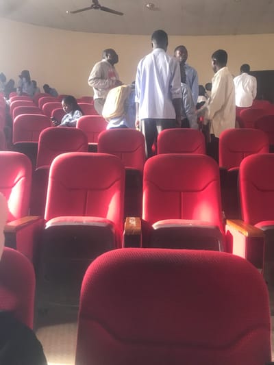 The image captures an interior view of an auditorium or lecture hall, oriented sideways, depicting rows of red upholstered seating. When viewed upright, a group of people, predominantly men, are visible on the left side of the frame, either seated or standing in the aisles between the rows of chairs. They are seen mostly from their backs or side profiles.
The subjects wear a mix of attire, including light-colored shirts and some traditional garments. One person in the mid-ground wears a distinctive yellow backpack. Another individual further back appears to be looking down. The setting is clearly an indoor venue designed for gatherings, characterized by the uniform red seating arranged in rows. A light-colored wall forms the background, and a dark ceiling fan is visible in the upper left corner of the upright image.
Bright natural light streams into the space from the top-left (when upright), illuminating the seats and creating distinct highlights and shadows, indicating it is daytime. The activity suggests an event, lecture, or gathering is either underway or about to commence, with attendees settling into or moving around the seating area.
Notable details include the sturdy red seats with dark armrests, some of which feature small labels. In one of the provided close-up crops, the text "NO SMOKING" is discernible on one of these labels, indicating a rule of the venue. The overall mood appears calm and organized. The location context indicates Durumi, Nigeria.