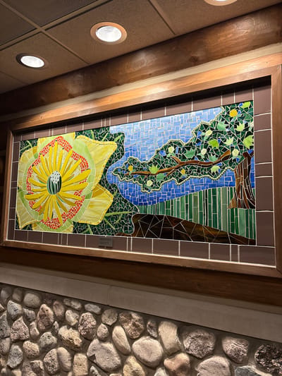 The image shows a large, colorful mosaic artwork framed by dark wood and mounted on a wall above a rustic stone facade. The mosaic depicts a stylized natural scene with a prominent yellow flower on the left, a tree with green leaves on the right, and a blue sky with some green foliage in the background. The mosaic is composed of numerous small, irregular pieces of colored glass or tile. A small plaque is visible at the bottom center of the mosaic frame. The ceiling above the artwork has acoustic tiles and recessed lighting. The stone facade below the artwork is made of rounded, rough-textured stones of various sizes and colors, primarily in shades of grey and brown. This mosaic is located in the New Harmony Inn, in New Harmony, Indiana, United States.