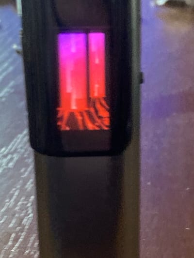 A close-up, slightly blurry, horizontal shot shows the display of a black electronic device, possibly a fitness tracker or e-reader. The display is illuminated with bright, vertical bars of light in shades of pink and purple, suggesting a screen that is on. The background is out of focus and appears to be a patterned surface, possibly a table or countertop, with a purple-toned light reflecting on it. The image is taken from a slightly elevated angle. There is no indication of the location within Chaiky, Ukraine.