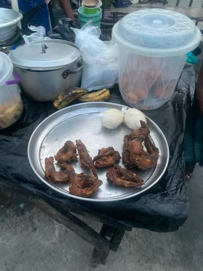 This image captures a street food stall in Iwofe, Nigeria, displaying various food items for sale. The scene is viewed from above, looking down at a makeshift setup.

The primary focus is a large, round, silver metal tray holding several pieces of fried meat, likely chicken or fish, characterized by a dark, crispy appearance. Beside these, on the same tray, are two small, round, white dough-like items, possibly puff-puff or similar fried snacks.

To the left of the tray, a large, transparent plastic container with a white lid is visible, filled with numerous orange-brown, roundish items, which also appear to be fried dough or snacks. Below this container, a bunch of ripe and semi-ripe bananas rests on the dark surface. A silver metal pot with a lid and handle is positioned further left, suggesting it holds other food items or ingredients. Various other plastic bags and smaller containers are scattered around the area, indicative of a vendor's typical setup.

The food items are arranged on a dark, possibly black, tarp or cloth that covers a surface, likely a table or stall. Underneath this, rough wooden planks are visible, forming the base of the stall. The ground surrounding the stall appears to be concrete or paved. A human arm and foot are faintly visible in the periphery, suggesting the presence of a vendor or customer.

The image depicts a common street food vending scene, highlighting local cuisine and an informal marketplace setting. No specific time of day, weather conditions, or emotions can be determined from the photograph, and there is no visible text.