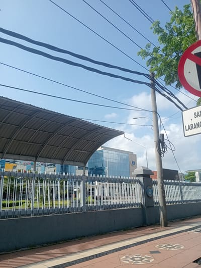 The image shows a clear sky over a city street scene in Surabaya, East Java. Overhead power lines crisscross the frame against the blue sky. A metal awning covers a walkway or seating area on the left. A tall, grey pole stands in the center, with a traffic sign on the right that appears to indicate "No Parking" or a similar restriction. Beyond a white metal fence, a modern building with blue glass windows is visible, along with some greenery. The ground in the foreground is paved with reddish-brown tiles and has some decorative circular inlays. A dark grey wall runs along the base of the fence. The overall scene is one of an urban environment under daylight.