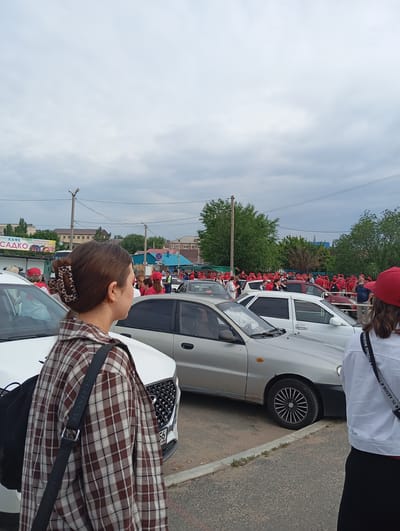 This is a medium shot, full-frame, eye-level outdoor photograph taken on a cloudy day in Sol-Iletsk, Russia. The foreground features a woman with brown hair pulled back with a leopard-print clip, wearing a brown and white plaid shirt and a black backpack slung over her shoulder. To her left, a white car is parked. In the background, a large group of people, mostly wearing red caps and shirts, are gathered in an outdoor area with several parked cars, including silver and red vehicles. A sign for a cafe is visible on a building in the background. The environment suggests a public gathering or event.