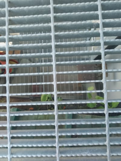 The image displays a close-up view of a metal grate. The grate is made of parallel metal bars, creating a grid pattern. Behind the grate, a blurry, out-of-focus scene is visible, including what appears to be some greenery and possibly some man-made structures. The lighting suggests an outdoor environment. The specific location in the city of Nicorești, Romania, cannot be determined from the visual information provided in the image.