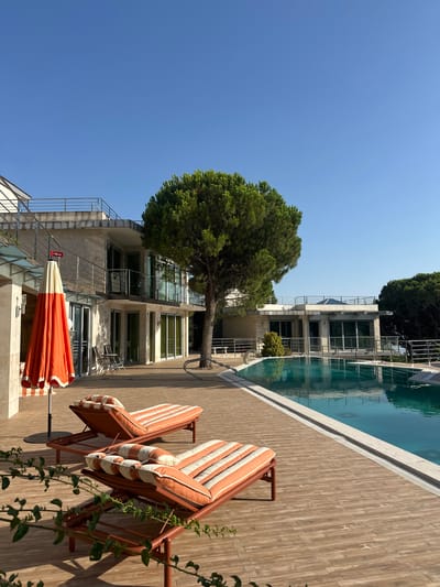The image depicts a serene outdoor scene at a luxury property in Brca, Montenegro. In the foreground, two orange and white striped chaise lounges with matching cushions are placed on a wooden deck. An orange and white beach umbrella is positioned to the left of the lounges, partially folded. To the right, a swimming pool with clear blue water reflects the bright sky and surrounding architecture.
The architecture features modern design with clean lines, concrete facades, and large glass windows. A multi-story building with balconies and railings is visible behind the chaise lounges, suggesting a residential or hotel setting. A tall, mature pine tree stands prominently in the middle ground, adding a natural element to the scene. The background reveals more of the property and dense green trees, hinting at a lush, possibly mountainous or coastal, environment typical of Montenegro. The sky is clear and blue, indicating fair weather. No people are present in the image.