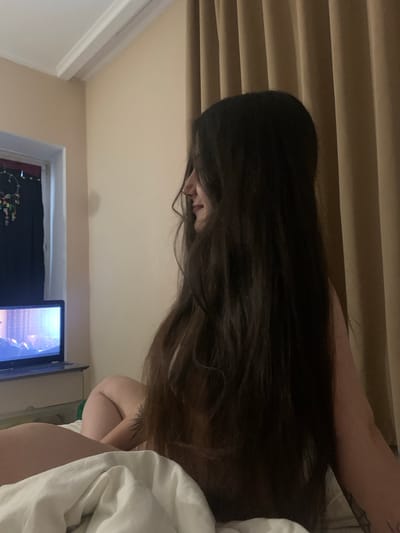 The image shows a woman with long dark hair lying in bed, facing away from the camera. She is looking at a laptop computer, which is open on the bed in front of her. The room is dimly lit, with beige curtains in the background and a window to the left. There is a dreamcatcher hanging on the window. The woman appears to be in a hotel room or a bedroom. The image does not provide any specific information about the location in the city of Сонячне, Ukraine.