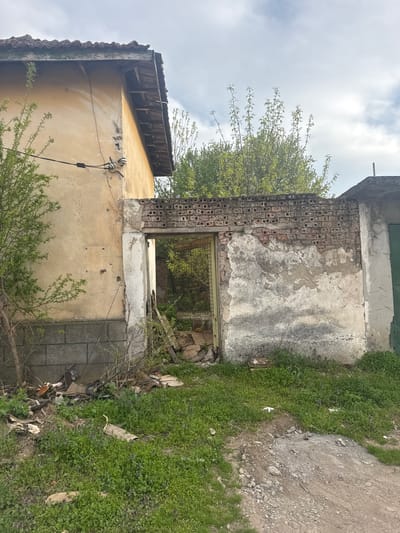 The image depicts a derelict building in what appears to be Gromshin, Bulgaria. The building has crumbling walls, with sections of brick and plaster missing. Vegetation is growing both on the roof and within the walls, indicating a state of advanced disrepair. The surrounding area is overgrown with weeds and appears unkempt. There are no people visible. The scene suggests abandonment and neglect.