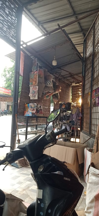 A black motorcycle is parked in front of a stall with a corrugated metal roof. The stall is cluttered with various items, including bags of goods, shelves filled with products, and what appears to be a broom leaning against a metal support. Behind the stall, a wire fence encloses an area where a chair and some folded fabric can be seen. Sunlight is streaming into the scene from the right, creating a warm glow. The ground is tiled. The time of day appears to be late afternoon or early evening, judging by the angle of the sun. The setting seems to be an outdoor market or roadside shop in Gampongteungoh, Indonesia. A sign in the background reads "Central Coffee".