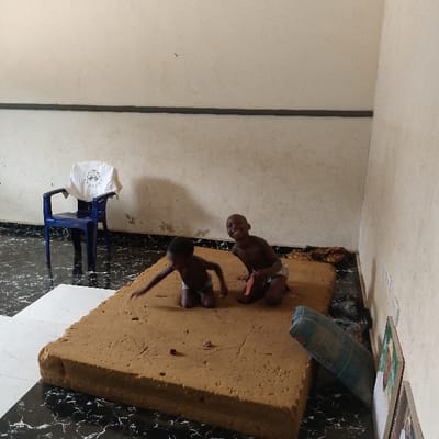 Two young children are playing on a large, brown mattress in a room with white walls. The room has a black and white tiled floor and a blue plastic chair with a white cloth draped over it. The room is located in Adanisievbe, Nigeria, at coordinates 5.953 latitude and 5.705 longitude. The children are smiling and appear to be having fun. One child is wearing a blue and white shirt, while the other is wearing a white undershirt. The children are seated on the mattress, and there is a small red toy on the floor near them.