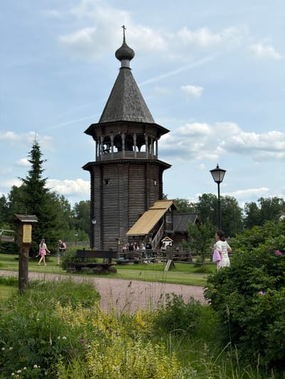 The image captures a scene in Nevskii Parkleskhoz, Russia, featuring a tall, multi-tiered wooden bell tower as the central architectural element. The bell tower has a conical roof topped with a cross and exhibits intricate wooden carvings. In the foreground, a winding gravel path leads through lush green grass and flowering plants, with a dense hedge of rose bushes to the right. Several people are visible: a woman in a pink skirt and a man in a dark shirt walk on the path near a wooden information board. Further back, a group of people are seated at picnic tables near a wooden structure with a thatched roof. A tall evergreen tree stands to the left of the bell tower. A lamppost is positioned on the right side of the image. The sky is partly cloudy with a blue hue.