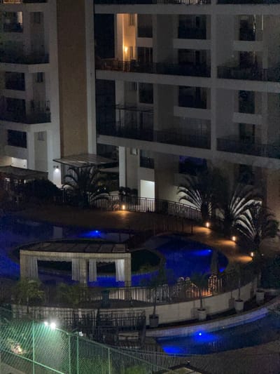 The image depicts an elevated perspective of a residential complex at night. A multi-story building with visible balconies is on the left. Adjacent is a swimming pool with blue illumination, surrounded by landscaping including palm trees and a wire fence. The specific location in Brasília, Brazil cannot be definitively determined from the image alone. No people or actions are visible.