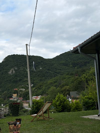 The image depicts a view from a residential property in the Golubac Municipality, Serbia, looking towards a heavily wooded mountain. In the foreground, a green lawn is visible with a lounge chair and two bags of charcoal. A telephone pole with a satellite dish stands prominently on the left side of the frame. In the middle ground, several houses with red-tiled roofs are nestled among the trees on the lower slopes of the mountain. The mountain itself is densely covered with dark green foliage, with some rocky outcrops visible on its upper reaches. The sky is overcast with gray clouds. The overall impression is of a semi-rural setting with a focus on nature and residential structures.