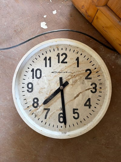 A round, vintage wall clock with a white face and black Arabic numerals is lying on a wooden floor covered in dust and debris. The clock face is stained and discolored, suggesting age and neglect. The hands of the clock are black, with the hour hand pointing to approximately 8:40 and the minute hand pointing to the 8. A black electrical cord runs across the top left of the image, connecting to the clock. In the upper right corner, a portion of a wooden structure, possibly a door frame, is visible. The overall scene suggests an abandoned or disused space. The location is Vazhiny, Russia.