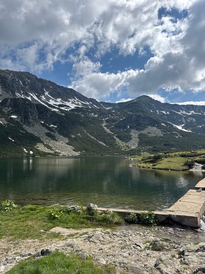 The image depicts a serene mountain lake scene in Avramovo, Bulgaria. Dominating the background are imposing, rugged mountains, partially covered with patches of snow, indicative of high altitude terrain. The slopes of the mountains are adorned with dark green coniferous trees, interspersed with rocky screes. A vast, calm lake occupies the mid-ground, its surface reflecting the sky and surrounding landscape. The water appears clear and deep, with visible ripples indicating a gentle breeze. In the foreground, a rocky shoreline transitions to grassy patches. A man-made concrete structure, possibly a small dam or pier, juts out into the water from the right side of the frame. The sky above is a mix of bright blue and scattered white clouds, suggesting fair weather. The overall impression is one of natural beauty and tranquility. There are no people visible in the image.