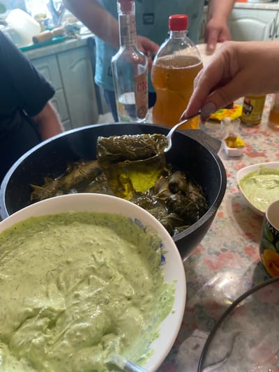 The image depicts a table laden with food, including what appears to be dolmades in a pot and a creamy green dip in a bowl. A person's hand is visible, using a spoon to scoop the dolmades. In the background, bottles of alcohol and a drink are present, along with glimpses of people. The overall setting suggests a meal or gathering. The image is set in the city of Shiryaevo, Russia.