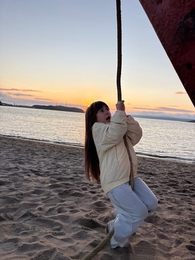 A young girl with long brown hair is holding onto a thick rope, appearing to swing or climb. She is wearing a light yellow jacket and light blue jeans. Her mouth is open in surprise or excitement. The rope is attached to a large red object on the right side of the frame, possibly part of a structure or vessel. The girl is standing on a sandy beach. In the background, a calm body of water stretches to the horizon, with a silhouetted landmass and some structures visible in the distance. The sky displays a soft sunset with hues of orange, yellow, and pink. The scene is set at sunset on a beach in Teriberka, Russia, a coastal village on the Kola Peninsula known for its dramatic Arctic landscapes and proximity to the Barents Sea.