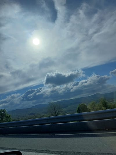 The image depicts a view from a moving vehicle, likely on a highway. The right side of the frame shows a gray metal guardrail and a portion of the asphalt road. Beyond the guardrail is a landscape of rolling hills covered in green vegetation, potentially trees. In the distance, there are faint outlines of mountains. The sky occupies the majority of the frame and is filled with various types of clouds, ranging from thin, wispy cirrus clouds to thicker, more defined cumulus clouds. The sun is visible through the clouds, appearing as a bright, white orb. Given the landscape and the general appearance, it's plausible that the image was taken in the vicinity of Lamego, Portugal, a region known for its mountainous terrain and scenic drives.