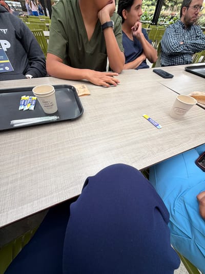 This image depicts a casual gathering of at least six individuals around a light wood-grain table, likely in an outdoor or semi-outdoor cafe or restaurant setting. The shot is taken from a slightly elevated, diagonal perspective, looking down at the table.
Several people are visible: a man in a blue plaid shirt and glasses sits with his arms crossed, looking forward. Next to him, another man in a dark blue t-shirt has his hand near his chin. Further down, a person in an olive green top, wearing a watch, rests their hands on the table. A person in a dark grey hoodie is partially visible in the foreground, bottom left. On the right side, parts of two other individuals are seen, one in a light blue top (possibly scrubs) and another in a dark blue garment. Green chairs and lush foliage in the background indicate a daytime scene.
On the table, there are various items including a black smartphone and a single paper cup. A black serving tray holds another paper cup, several small yellow and blue packets, and straws. A brown paper bag is also present.
Visible text on the small packets and one of the paper cups includes "Incauca." Another paper cup on the tray reads "ESTE VASO ES LIBRE DE ARBOLES" (This cup is free of trees) and "Lining." Partial text "P", "ACIÓN", "Y LED", and "PUON" is visible on the hoodie of the person in the foreground. The presence of "Incauca" suggests a brand commonly found in Colombia. The scene conveys a relaxed atmosphere, possibly a coffee break or an informal meeting.