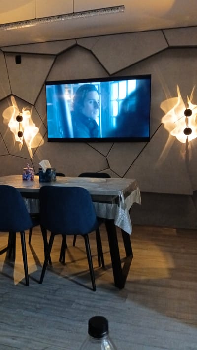The image depicts an indoor scene, likely a living room or dining area, featuring a wall-mounted television displaying a scene from a movie or show. The wall itself has a geometric, textured design. Two decorative wall sconces with a warm, diffused light are visible on either side of the television. In the foreground, a dining table is covered with a transparent plastic tablecloth. On the table, there is a box of tissues and a decorative jar. Two dark blue chairs are positioned around the table. The floor is made of light-colored wood planks. A partially visible plastic bottle with a black cap is in the lower right corner of the image. The scene is well-lit, with light emanating from the sconces and the television. There are no people directly visible in the frame, and no specific geographical indicators point to Gulkevichi, Russia.