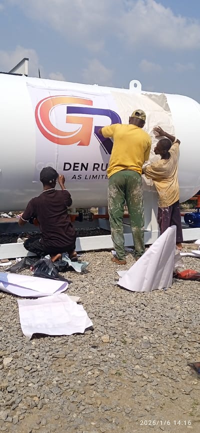 On January 6, 2026, at 14:16 local time, in Isheri-North, Nigeria, three males are engaged in applying a large commercial decal to a horizontal, white cylindrical storage tank. The tank, positioned outdoors on a gravel and dirt surface, rests on a dark-colored support structure atop a concrete base.
The decal, partially adhered to the tank, features an orange and purple "GR" logo prominently at the top, with visible text "DEN RU" followed by "GAS LIMITED" below, indicating the company "GOLDEN RUGAS LIMITED."
One male, wearing a dark brown short-sleeved top and a black baseball cap, kneels on the ground, barefoot, manipulating a lower section of the decal. Surrounding him are crumpled black plastic and white paper debris. A second male, clad in a bright yellow short-sleeved t-shirt, a camouflage baseball cap, and paint-stained green trousers, stands upright, smoothing the upper-center portion of the decal with both hands. He wears light brown sandals. The third male, wearing a light yellow short-sleeved top and dark trousers (possibly stained), stands adjacent to the second, assisting in smoothing the upper-right section of the decal. His feet are bare.
The immediate ground area consists of coarse gray gravel mixed with dirt and scattered with discarded white paper remnants from the decal application. The scene is illuminated by bright daylight under a partially cloudy blue sky. A building with a dark roof is visible in the far-left background.