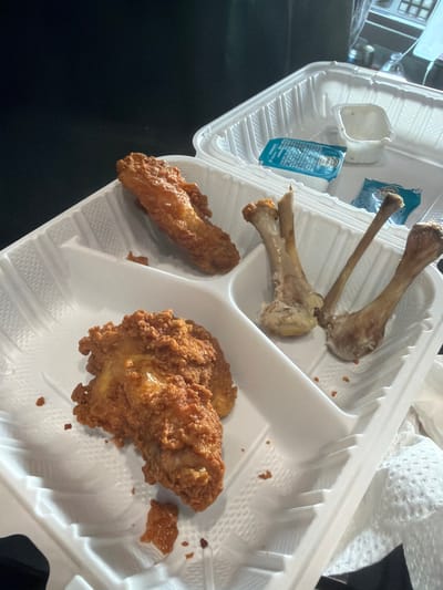 A close-up, overhead shot shows a white styrofoam takeout container filled with leftover fried chicken and bones. Two pieces of fried chicken, one larger and more intact than the other, are positioned in the lower left section of the container. Three chicken bones, scraped clean of meat, are arranged in the upper right section. Two small, foil-wrapped condiment packets, one blue and one white, and a small, empty plastic dipping sauce cup are visible in the upper right compartment of the container. The container itself is divided into three sections. The background is dark and out of focus, with hints of a car interior, including a dashboard and a steering wheel, suggesting the food may have been consumed in a vehicle. The image has a slightly grainy texture and is lit from above, casting subtle shadows. There is no information to suggest the location in the city of Raphine, United States.