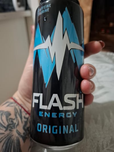 A person's hand is holding a black energy drink can with a lightning bolt design. The can has text that reads "FLASH ENERGY ORIGINAL" and "CAFFEINE TAURINE". The person has tattoos on their arm and is wearing a red string bracelet. The background is blurred, showing what appears to be a bed or sofa and some furniture. There is no specific indication of the location being in Novlyanka, Russia.