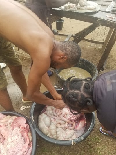 Two people are processing animal innards in a muddy outdoor area. The man on the left is shirtless and wearing shorts and is reaching into a black bin of animal intestines. The woman on the right is leaning into the bin, her hair in braids and wearing a black shirt, while holding the intestines with her right hand.  Another man is visible in the background, standing on a wooden table with a small black bucket and a white-and-grey object. The coordinates provided, latitude 7.81 and longitude 6.704, place the scene in Pati Ridge, Nigeria.