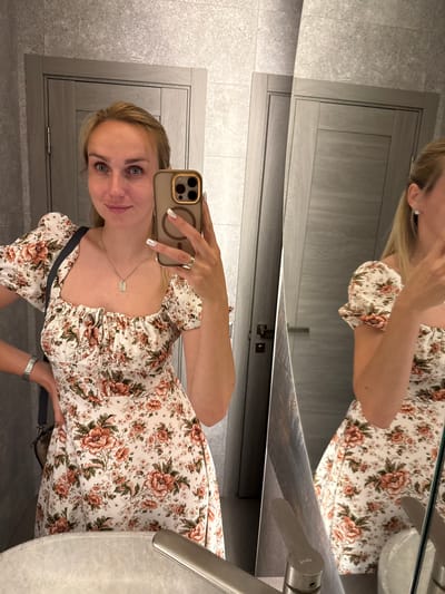 The image shows a young woman taking a mirror selfie. She is wearing a floral print dress with short puffy sleeves. She has blonde hair and is smiling at the camera. She is holding a smartphone in her right hand, which is angled to capture her reflection. A silver necklace with a rectangular pendant is visible around her neck. On her left hand, she is wearing a ring. The background appears to be a bathroom, with a large mirror and textured gray walls. In the reflection, part of a sink and faucet are visible. The lighting suggests it could be daytime or well-lit artificial light. The woman's expression is pleasant and she seems to be posing for the photo. There is no visible text in the image.