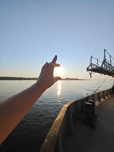 The image captures a hand reaching out towards the setting sun over a wide river, with the deck of a ship in the foreground. The sun is a bright orb reflecting on the water. A person's arm and hand are positioned in the lower left portion of the frame, with fingers partially obscuring the sun. The river stretches across the mid-ground, with a distant tree line on the far bank. The ship's deck, made of dark material with metal railings and bollards, occupies the right and bottom portions of the image. A metal walkway or platform extends from the ship towards the right. The sky is clear and blue, indicative of fair weather. The location is not identifiable as Lesosibirsk, Russia, as there are no distinguishing landmarks or context provided in the image itself.