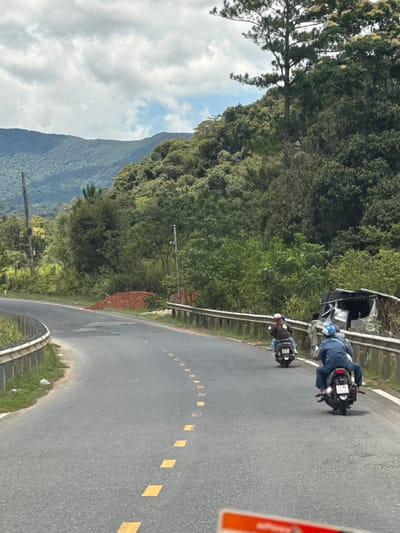 The image shows a winding asphalt road in a rural, mountainous area. On the right side of the road, there is a guardrail, and beyond that, a dense forest covers the hillside. The sky is cloudy with patches of blue. In the foreground, two motorbikes are traveling away from the viewer. The first motorbike has a rider wearing a blue jacket and helmet, and the second motorbike, slightly ahead, has a rider in a brown jacket and a white helmet. The road has yellow dashed lines marking the lanes. On the left side of the road, there is another guardrail and a grassy embankment. A pile of reddish-brown soil is visible on the side of the road near the center of the image, and to the right of that, a makeshift shelter made of corrugated metal and tarps is leaning against the guardrail. The scene appears to be a typical roadside in a mountainous region of Vietnam, potentially near Đưng K'Si.