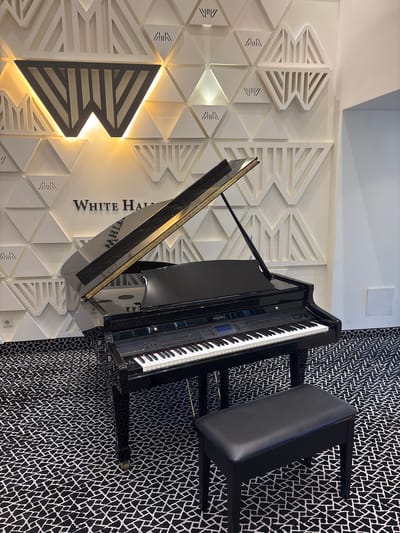 An upright grand piano with an open lid and a black bench sits on a black and white geometric patterned carpet in front of a white wall with a honeycomb-like geometric design and the text "WHITE HALL" in black lettering. The wall is illuminated with warm lighting, highlighting a stylized "W" logo. The piano has a black glossy finish and visible keys and control panel, with the brand name "SUZUKI" clearly visible. The scene is well-lit and appears to be indoors, likely a performance space or a lobby. The city of خربیه السوق, Jordan is not discernible from the image itself.