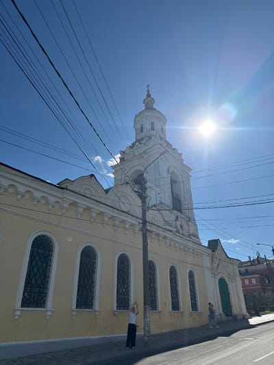 In the city of Yelets, Russia, a low-angle shot captures a pale yellow building with arched windows and white decorative elements. To the right, a white bell tower with arched openings and a cross at its apex rises against a bright blue sky. The sun is prominently visible, creating a lens flare. Overhead, a network of dark electrical wires crisscrosses the frame. On the sidewalk in front of the building, a person in dark pants and a light shirt stands facing away from the camera, with one arm raised. Further to the right, two people, one holding a child, are visible near a bright green door. The scene includes a paved street in the foreground.
