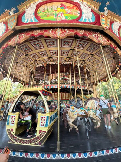 A carousel is a popular amusement ride consisting of a rotating circular platform with seats for riders, usually in the form of horses or other animals, attached to poles. The carousel in this image features ornate decorations, including colorful paintings and gold accents, typical of traditional carousels. The subjects include several people, some of whom are children, enjoying the ride. They are seated on the carousel's horses, some appearing to be engaged in conversation, while others seem to be simply enjoying the experience. The setting appears to be an outdoor amusement park, likely in Castelnuovo del Garda, Italy, as suggested by the provided location context. The background shows trees and greenery, indicating a pleasant, possibly sunny day, suggesting the activity is taking place during daylight hours. The mood is likely joyful and celebratory, as is common with amusement park rides. The carousel itself is a complex structure with a detailed wooden ceiling and supporting poles, all contributing to its vintage charm. One of the carousel cars has the number "2" visible on it.