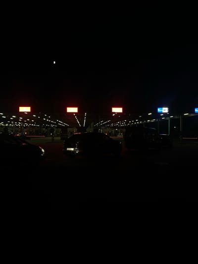 The image shows a nighttime scene, possibly at a border crossing or a parking area, with a row of lights and illuminated signs. The setting appears to be outdoors, with a dark sky and what looks like a distant moon. There are several vehicles, including a dark-colored car with visible license plate text, and silhouettes of people. Red and blue rectangular signs are visible, as well as a string of small white lights that illuminate a pathway or area. The overall impression is one of activity and order in a dimly lit environment, suggesting a functional area operating at night. The location context of Terespol, Poland, could imply a border crossing or transportation hub. The image is oriented vertically, with the lights and signs extending upwards from the bottom.
