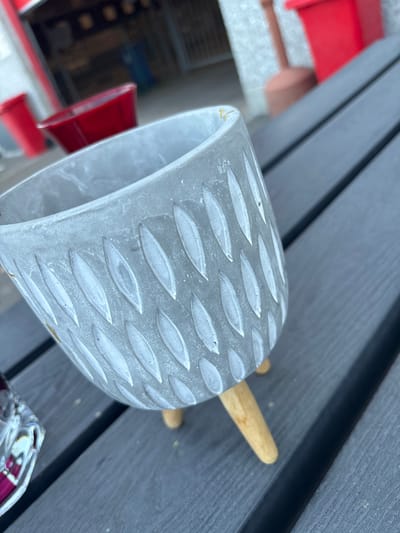 The image shows a close-up of a light gray, textured pot with three wooden legs, placed on a dark gray wooden surface. The pot has a pattern of elongated, leaf-like indentations filled with a lighter gray substance. In the background, out of focus, are several red planters and a section of a building with a textured facade. The scene appears to be outdoors, possibly on a patio or deck. There are no people visible in the image, and no specific location within the city of Boom, Belgium, can be identified from the visual cues.