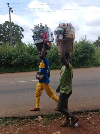 In Ugwolawo, Nigeria, two men walk from right to left along the shoulder of a paved road under an overcast sky. The man in the foreground on the left wears a blue and yellow short-sleeved shirt, yellow trousers, and red flip-flops, with a dark shoulder bag slung across his chest. He balances a large, dark container on his head, filled with various packaged consumer goods secured by white strapping. His left arm is raised, steadying the load. The man in the background on the right wears a green and white striped short-sleeved shirt, dark trousers, and dark slide sandals. He carries a light-brown plastic basket on his head, similarly packed with various items and secured with white strapping, with his left arm also raised for support. The road surface is dark asphalt with a faint white line visible. The immediate roadside consists of a narrow strip of lighter pavement and reddish-brown dirt with scattered dry leaves and sparse ground vegetation. Dense green foliage and trees line the background. A utility pole with overhead wires is visible in the upper left, behind the trees.