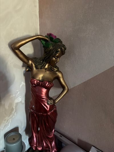 The media file depicts a close-up, horizontal shot of a decorative figurine of a woman lying down. The figurine is predominantly gold, with accents of red and green. The woman is adorned with a flowing, metallic red dress that drapes around her body. Her hair is long and dark, with floral elements in shades of pink and green adorning her head. She is resting on her left side, with her left arm bent and her head supported by her hand. Her right arm is bent at the elbow and rests on her hip. The figurine is placed on a white, textured surface that appears to be fabric or a similar material. In the background, there is a textured wall with a diagonal shadow cast across it, suggesting a light source from the right. To the far right of the frame, a portion of a grey cylindrical object, possibly a candle, is visible. The lighting appears to be artificial and creates some glare on the figurine's metallic surfaces. There is no visible text. The scene is static, with the figurine being the sole subject. The overall impression is that of a decorative piece displayed in an indoor setting, possibly a home.