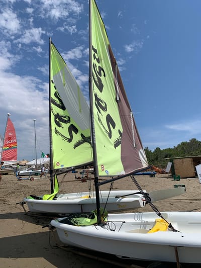 This image depicts a sunny day on a sandy beach, likely in Orbetello, Italy, given the presence of multiple sailboats and a beachside setting with trees and a small structure in the background. In the foreground, two white sailboats are resting on the sand, each with a mast and a sail. The sails are predominantly light green with dark lettering and some brown accents. One of the sails displays the text "#ZESTISBEST" in black. The boats appear to be small dinghies, possibly used for recreational sailing or training.
Behind these two boats, other sailboats are visible, including a red and white sailboat with the number "50319" and a rainbow-colored design on its sail. A few people are scattered on the beach, some appearing to be involved with the boats, while others are standing or walking around. The environment is characterized by sand, the sea or a large body of water, and a clear blue sky with scattered white clouds. In the mid-ground, a small wooden structure with a roof and an open front is situated, possibly a beach shack or storage area. The overall scene conveys a sense of leisure and outdoor activity.