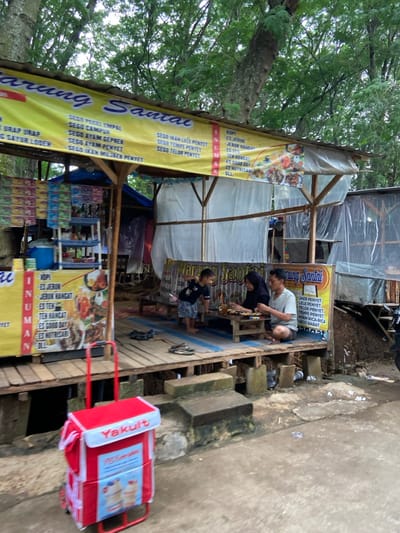A small, open-air eatery is bustling with activity as two adults and a child gather around a table. The structure is adorned with vibrant yellow banners listing menu items such as "Sego Ayam Penyet" and an array of refreshing beverages including "Es Jeruk". Stacked cartons of products and a red "Yakult" cooler on wheels suggest a variety of offerings for customers seeking a quick bite or refreshing drink. The scene captures a lively moment in a local food establishment.