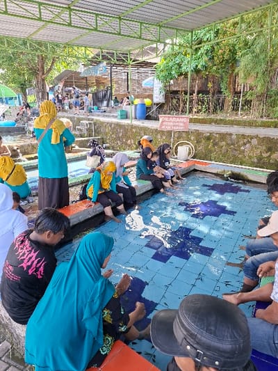 The image shows a group of people in a small, tiled pool. The pool is filled with clear water and there are small fish swimming around. The people are sitting in the pool with their feet in the water, allowing the fish to nibble on their skin. There are people watching from the side of the pool, and a sign in Indonesian can be seen that says "Dilarang Mengambil Ikan Tarap", meaning "It's forbidden to take the Tarap fish." The pool is located in Kecamatan Tulung, Indonesia, at latitude -7.592 and longitude 110.65. The people are all wearing traditional Indonesian clothing. The area is covered by a roof supported by metal beams. There are trees and buildings in the background. The scene is one of leisure and relaxation. There are also several small, plastic containers placed along the side of the pool. The pool is surrounded by a stone wall.