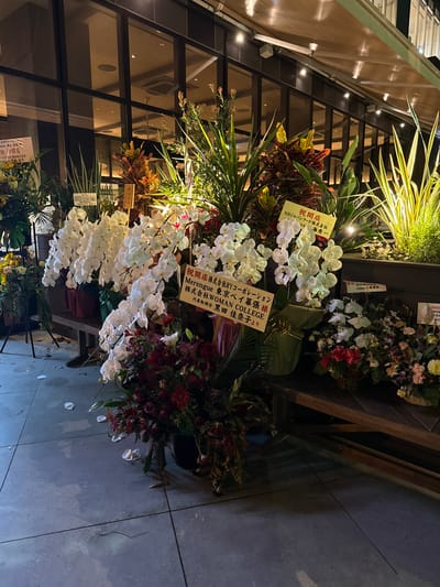 This is a horizontal, medium-shot, eye-level photograph of a storefront display of flower arrangements, likely for a grand opening, in the city of Chiba, Japan. The main subjects are several large, ornate floral displays, predominantly featuring white orchids and dark red flowers. In the background, a modern building with large windows and an awning is visible. The ground is a light-colored, tiled surface with some scattered flower petals.

To the left of the frame, a white sign with Japanese text reads "祝開店" (Shukaiten - Grand Opening) and lists corporate sponsors. Further to the left, a smaller arrangement with yellow flowers is partially visible. In the center, a prominent display of white orchids cascades downwards. To the right of the orchids, another yellow sign with Japanese text, "祝開店" (Shukaiten - Grand Opening), lists "NHジャパンフード株式会社" (NH Japan Food Co., Ltd.) and the name "佐藤 秀一" (Sato Shuichi).

Behind the main floral displays, there are tall, spiky green plants. To the right, other floral arrangements are visible, one in a woven basket with a red ribbon. Another sign with Japanese text, "祝開店" (Shukaiten - Grand Opening), is partially visible on the far right. The overall lighting suggests it is either dusk or evening, with artificial lights illuminating the scene from above and within the building. The environment appears to be an urban setting.