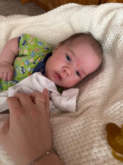 A close-up, high-angle shot captures a baby lying on a cream-colored, textured blanket. The baby is dressed in a green onesie with a pattern of cartoon cars and a white undershirt. The baby's head is turned slightly to the right, revealing blue eyes, a pinkish complexion with some redness on the forehead and cheeks, and fine dark hair. A person's hand, wearing a thin gold ring, is gently touching the baby's chest area. A portion of a tan pacifier is visible on the right side of the frame. The background shows the edge of wooden furniture. The image does not contain information about the city of Выльгорт, Russia.