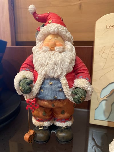 A vertically oriented, medium shot photograph features a figurine of Santa Claus displayed indoors. The figurine is detailed, with a red suit trimmed in white fur, a blue vest adorned with gold stars, and brown pants. Santa has a white beard and rosy cheeks, and he is holding a red and black checkered star. His hands are gloved in green. Behind the figurine, a light-colored object with the Cyrillic text "Лес" is visible. The background suggests an indoor setting, possibly a shelf or table, with a dark surface in the foreground and a wooden surface behind. The lighting appears to be artificial, and the overall mood is festive and decorative.