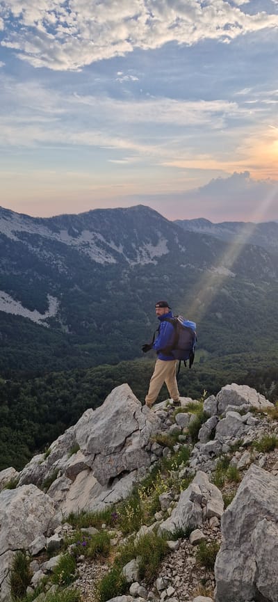 A man wearing a blue jacket, tan pants, a black baseball cap, and a large backpack stands on a rocky mountain peak. He is facing to his left, looking out over a vast valley filled with green trees. In the background, more mountains are visible, with a colorful sunset sky above. The foreground is dominated by large, gray rocks and scattered green grass with small purple and yellow wildflowers. The location is identified as Ubla, Bosnia and Herzegovina.