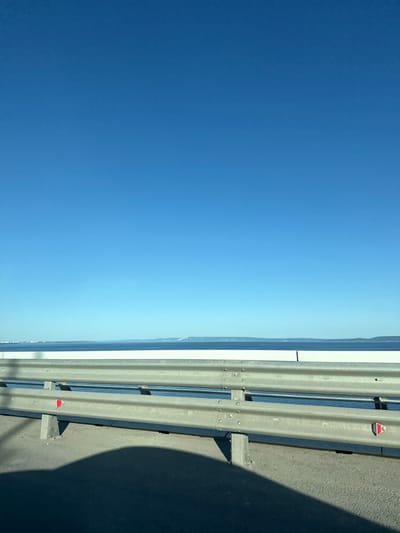 The image shows a view from a bridge in the city of Klimovka, Russia. The sky is a clear blue, with a slightly lighter blue near the horizon where it meets a distant shoreline. A metal guardrail runs along the right side of the image, next to a concrete surface. The guardrail has small red and white reflectors. A dark shadow is cast onto the concrete surface, likely from a vehicle or structure.