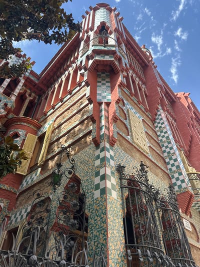 The image shows the exterior of Casa Vicens, located in Barcelona, Spain. It is a building with a reddish-pink brick facade, adorned with intricate decorative tiles in green, white, and yellow. The architecture features sharp angles, arched windows with green shutters, and ornate wrought-iron balconies. A tree with large, dark green leaves is visible in the upper left corner, casting shadows on the building. The sky above is a clear blue with scattered white clouds. The image is a low-angle shot, emphasizing the height and elaborate details of the building.